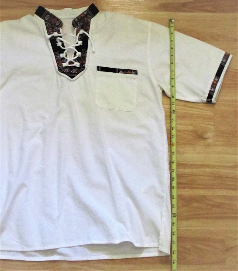 May include: White short-sleeved shirt with a lace-up neckline and a patterned trim. The shirt has a chest pocket with a patterned trim. The shirt is 28 inches long.