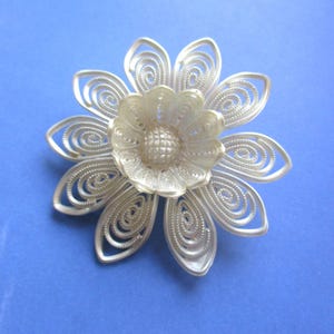 May include: A silver-toned flower brooch with intricate filigree detailing. The brooch features a central flower with a textured center, surrounded by delicate petals with spiral designs. The brooch is set against a blue background.