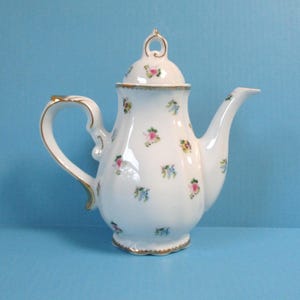May include: White porcelain teapot with a lid, handle, and spout. The teapot is decorated with small pink and blue floral patterns and gold trim. The handle and lid finial have gold accents. The teapot is on a blue background.