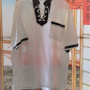 May include: White short-sleeved shirt with a black and white geometric pattern around the neckline. The shirt has a lace-up neckline and a small pocket on the right side.