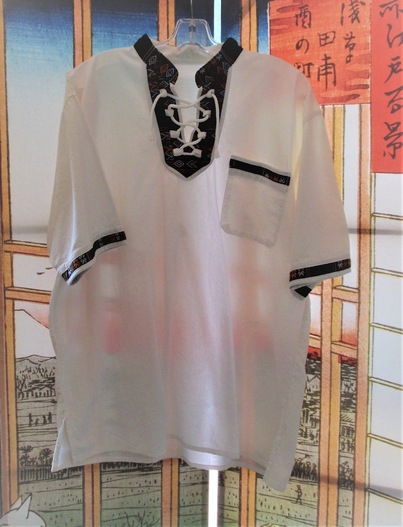 May include: White short-sleeved shirt with a black and white geometric pattern around the neckline. The shirt has a lace-up neckline and a small pocket on the right side.