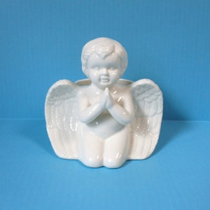 May include: A white ceramic angel figurine kneeling with hands clasped in prayer. The angel has detailed wings and is set against a blue background. The figurine is a decorative item.