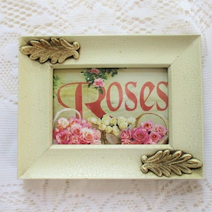 May include: A white frame with gold leaf accents, featuring a vintage-style print of pink roses with the word "Roses" in red lettering.