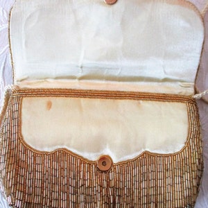 May include: A vintage gold beaded handbag with a scalloped flap and magnetic closure. The interior is a cream-colored satin. The bag has a long, thin, cream-colored cord strap. The exterior is covered in vertical gold beads.