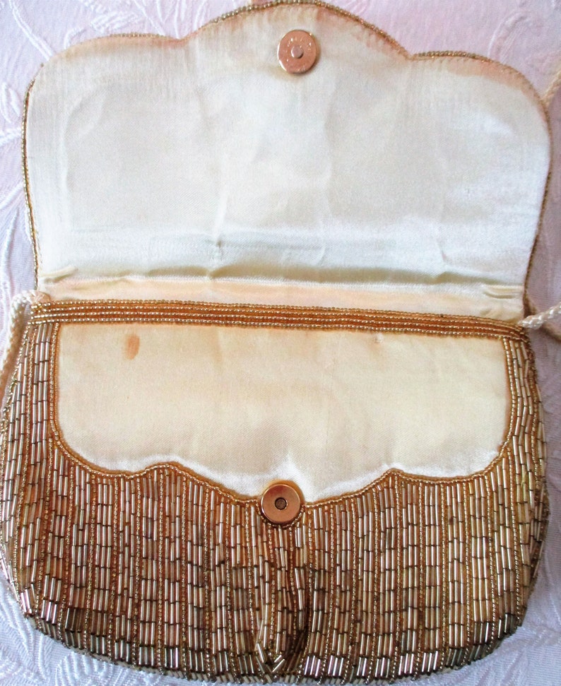 May include: A vintage gold beaded handbag with a scalloped flap and magnetic closure. The interior is a cream-colored satin. The bag has a long, thin, cream-colored cord strap. The exterior is covered in vertical gold beads.