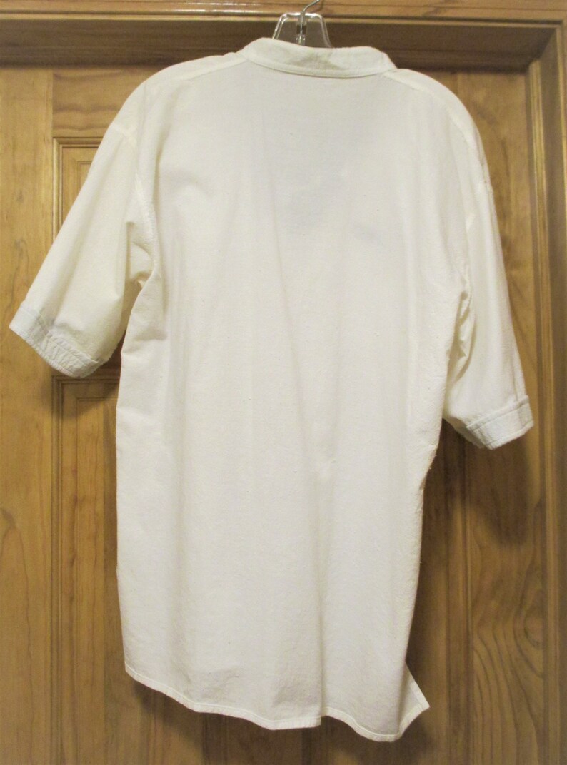 May include: A white short-sleeved button-down shirt hanging on a hanger. The shirt is made of a lightweight fabric and has a relaxed fit.