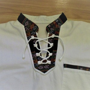 May include: White shirt with a black and colorful patterned collar and a lace-up neckline. The shirt has a pocket on the right side with a black and colorful patterned trim.