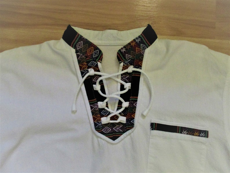 May include: White shirt with a black and colorful patterned collar and a lace-up neckline. The shirt has a pocket on the right side with a black and colorful patterned trim.
