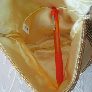 May include: Open view of a small, cream-colored beaded handbag with a gold trim. The interior is a light yellow satin, and an orange pen is inside. The bag has a small pocket and a tag.