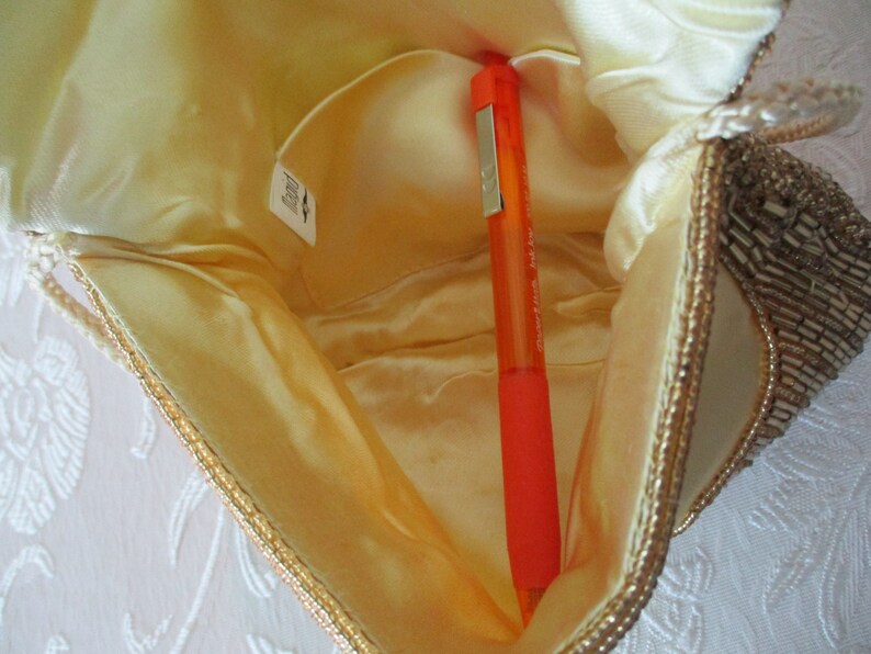 May include: Open view of a small, cream-colored beaded handbag with a gold trim. The interior is a light yellow satin, and an orange pen is inside. The bag has a small pocket and a tag.