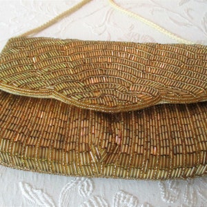 May include: Gold beaded clutch with a flap closure and a cream-colored shoulder strap. The rectangular bag is covered in small, vertical gold beads, creating a textured surface. The flap has a scalloped edge.