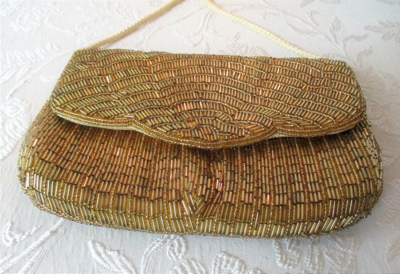 May include: Gold beaded clutch with a flap closure and a cream-colored shoulder strap. The rectangular bag is covered in small, vertical gold beads, creating a textured surface. The flap has a scalloped edge.