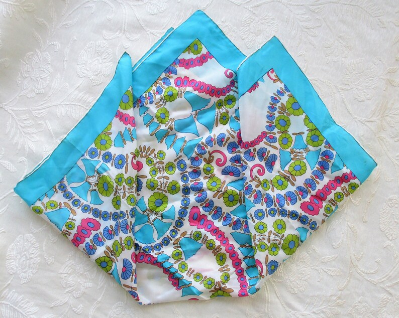 May include: A white silk scarf with a blue, green and pink floral pattern. The scarf has a turquoise border.