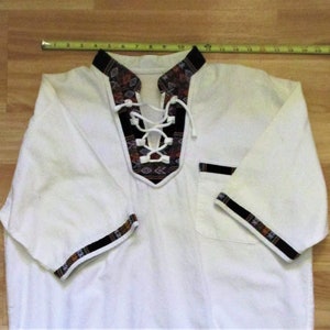 May include: White short-sleeved shirt with a lace-up neckline and black and brown patterned trim. The trim features geometric designs.