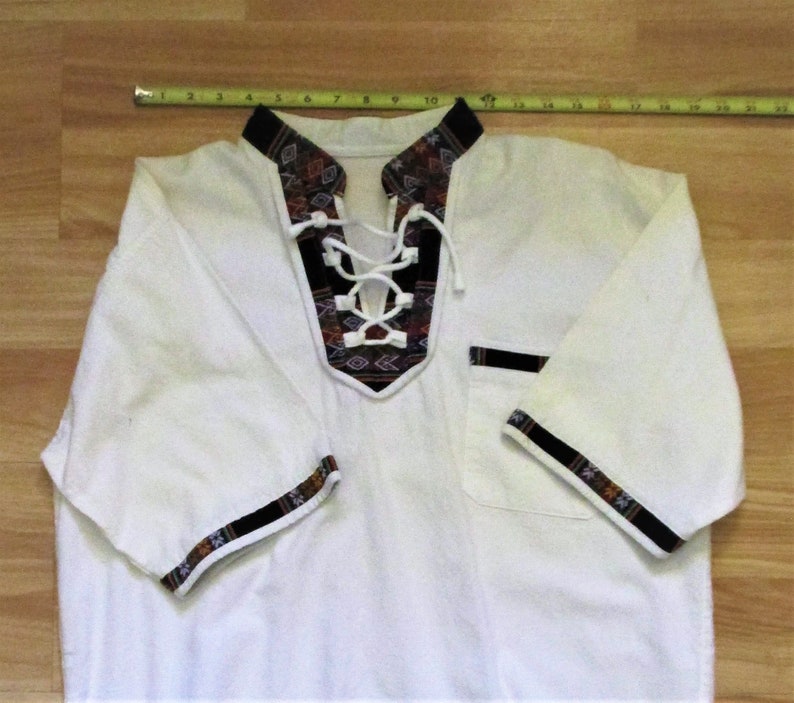 May include: White short-sleeved shirt with a lace-up neckline and black and brown patterned trim. The trim features geometric designs.