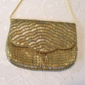 May include: A gold beaded clutch purse with a flap closure and a thin shoulder strap. The purse is covered in small, vertical beads in shades of gold and silver, creating a textured, wave-like pattern. The strap is a thin, woven cord.