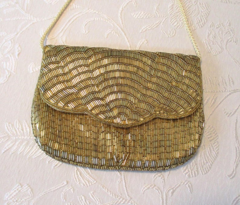 May include: A gold beaded clutch purse with a flap closure and a thin shoulder strap. The purse is covered in small, vertical beads in shades of gold and silver, creating a textured, wave-like pattern. The strap is a thin, woven cord.