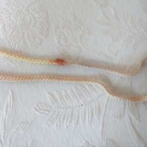 May include: A delicate, cream-colored necklace with small, orange-red beads. The necklace is made of a woven material and lies on a white, textured surface. The beads are evenly spaced along the necklace.