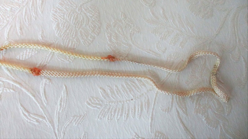 May include: A delicate, cream-colored necklace with small, orange-red beads. The necklace is made of a woven material and lies on a white, textured surface. The beads are evenly spaced along the necklace.