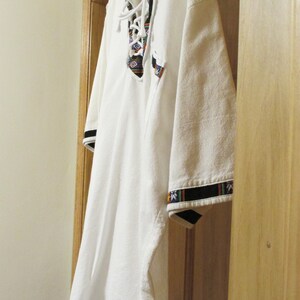 May include: White shirt with a colorful embroidered collar and black trim on the sleeves. The shirt is hanging on a hanger.