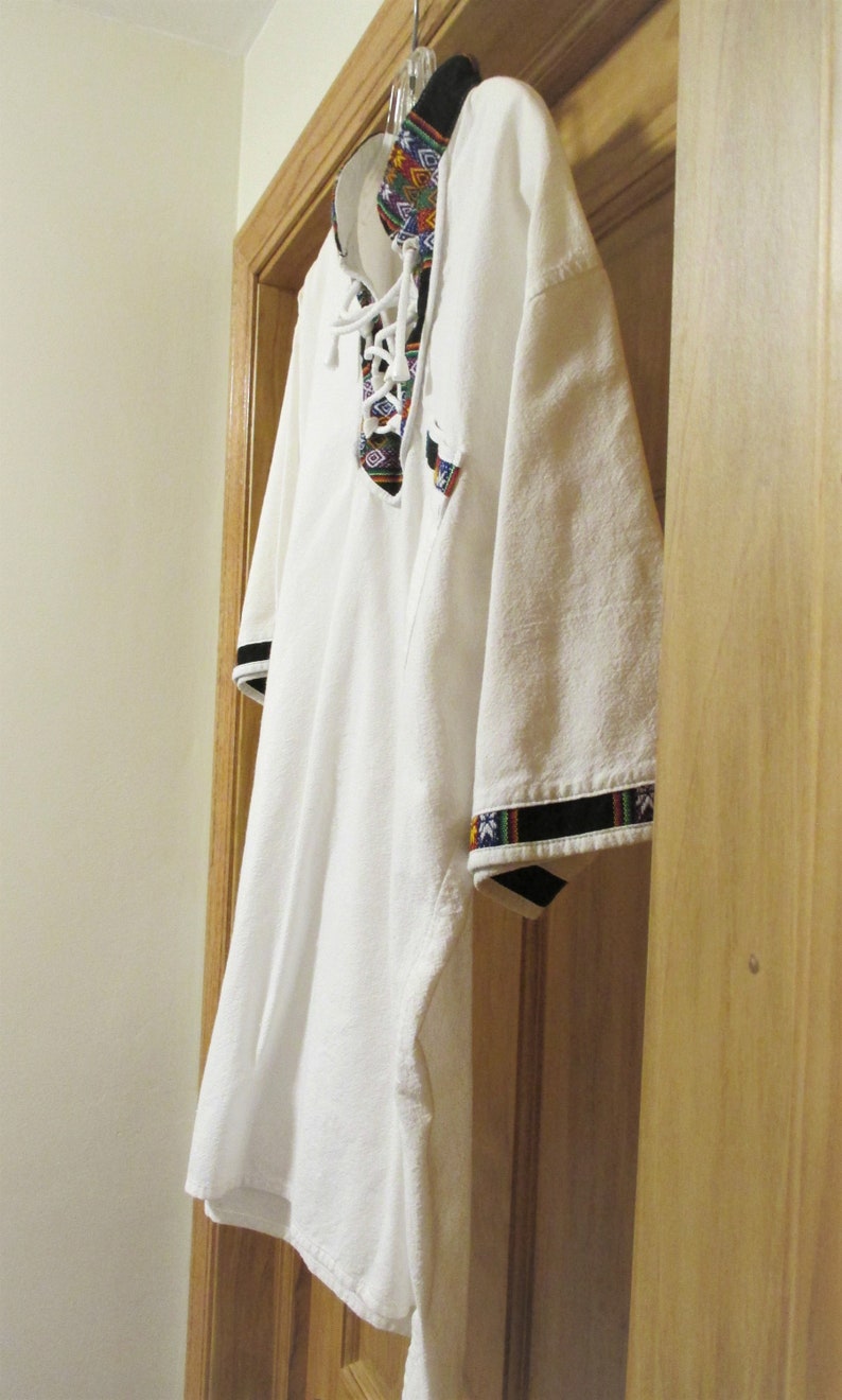 May include: White shirt with a colorful embroidered collar and black trim on the sleeves. The shirt is hanging on a hanger.