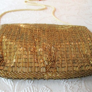 May include: A gold beaded clutch with a cream-colored shoulder strap. The rectangular bag is covered in vertical rows of small, golden beads. The strap is a twisted cord. The bag is on a white patterned surface.
