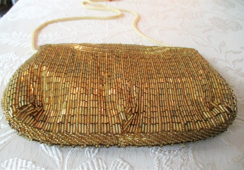 May include: A gold beaded clutch with a cream-colored shoulder strap. The rectangular bag is covered in vertical rows of small, golden beads. The strap is a twisted cord. The bag is on a white patterned surface.