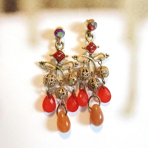 May include: A pair of gold-tone chandelier earrings featuring iridescent round studs, red square crystals, filigree metal spheres, and dangling teardrop-shaped beads in vibrant orange and translucent red hues suspended from metal loops.