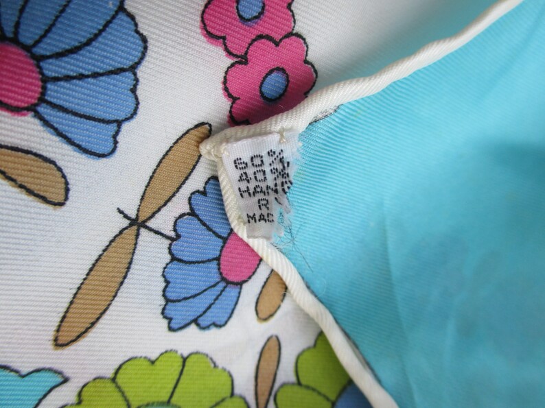 May include: Close-up of a white fabric with a blue, pink and green floral pattern. The fabric has a tag attached that reads "60% 40% HAND R MADE".