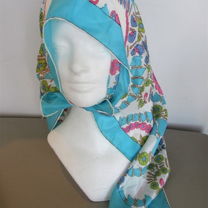 May include: A white, blue, pink and green floral patterned scarf with a turquoise blue border. The scarf is folded and draped over a white mannequin head.