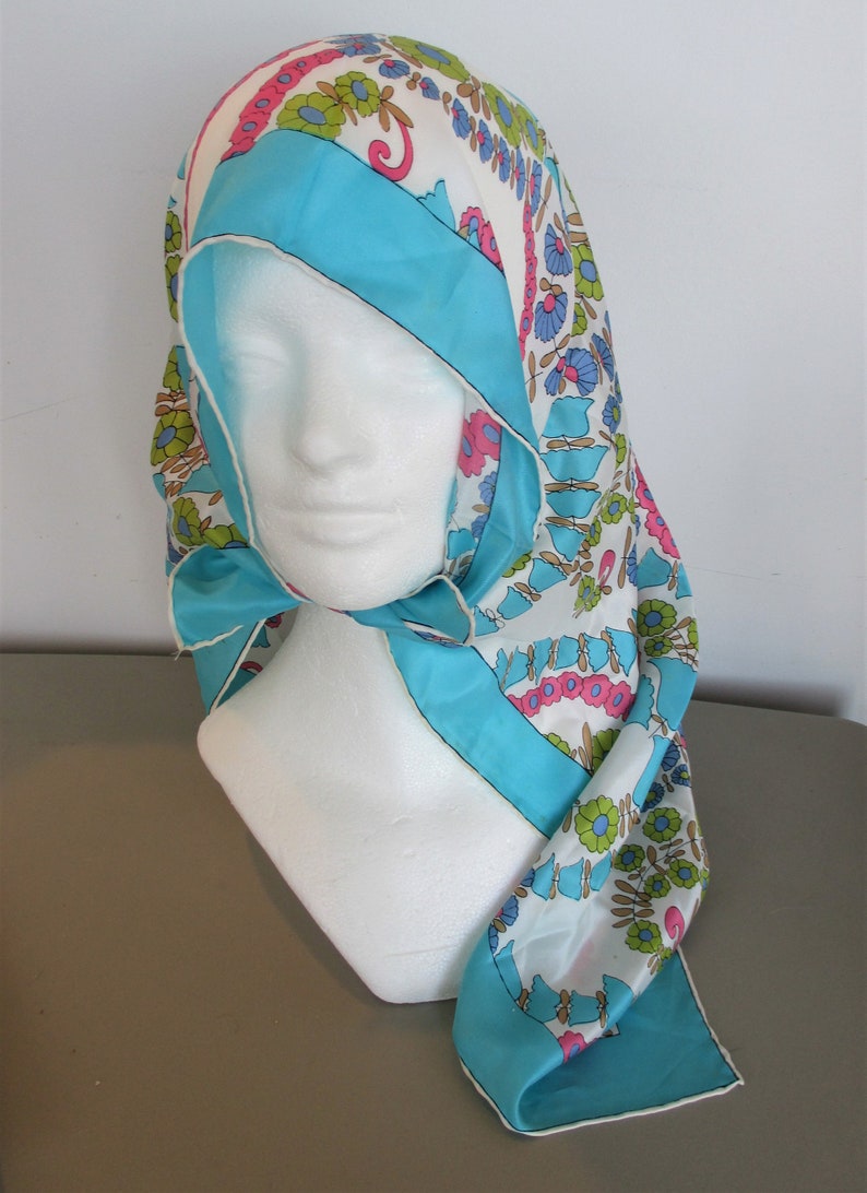 May include: A white, blue, pink and green floral patterned scarf with a turquoise blue border. The scarf is folded and draped over a white mannequin head.