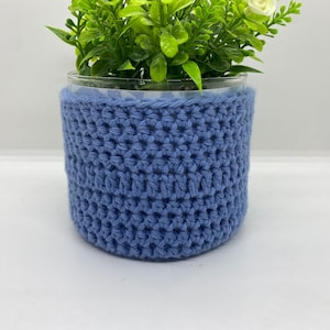 May include: A blue crocheted plant pot cover for a glass container. The pot cover is made with a simple stitch pattern and has a smooth, even texture.