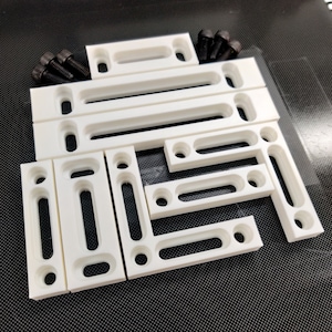 May include: A set of white plastic corner brackets with slots for screws. The brackets are arranged in a variety of configurations, with some stacked on top of others. There are also several black screws included in the set.