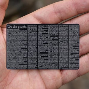 May include: A black rectangular card with the words "We the people" at the top. The card is filled with small text, resembling a document. The card is held in a person's hand.