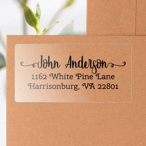 May include: Clear address label with black text. The label reads "John Anderson" in a script font, followed by the address "1162 White Pine Lane, Harrisonburg, VA 22801". The label is on a brown envelope.