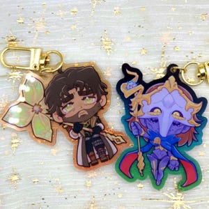 May include: Two acrylic keyrings with gold-coloured clasps. One keyring features a cartoon character with a beard, holding a staff. The other keyring depicts a character in a helmet and cape, holding a staff. Both keyrings have a glossy finish.