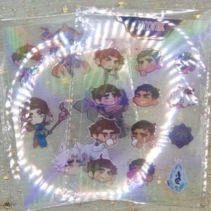 May include: Holographic sticker sheet with cartoon character designs. The stickers depict various characters with diverse hairstyles and clothing. Some stickers are shaped like gems or stars. The word "JAYVIK" is printed at the top of the sheet.