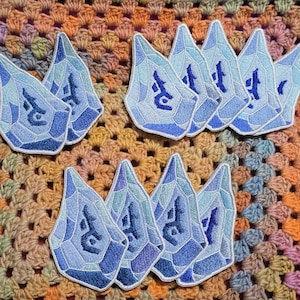 May include: A collection of blue crystal-shaped embroidered patches. Each patch features a geometric design with varying shades of blue and a dark blue symbol. The patches are arranged on a colourful crocheted background.