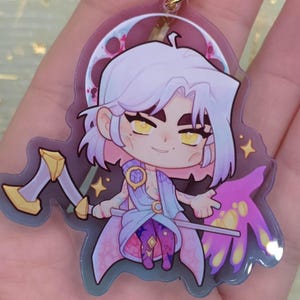 May include: A clear acrylic charm featuring a cartoon character with white hair, yellow eyes, and a light blue outfit. The charm has a gold-colored chain and a design that includes a crescent moon, stars, and a purple and pink wing-like shape.