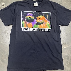 May include: Black t-shirt with a graphic of two Teenage Mutant Ninja Turtles. The text reads "Pizza Dude's Got 30 Seconds." The turtles are depicted in their iconic colors, with purple and orange masks.