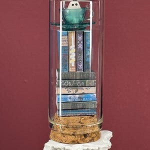 May include: A clear glass cylinder cloche with a cork base containing a stack of miniature books topped with a teal teacup and a small white ghost figure. The books feature various blue and grey spines with text including Book Magic.