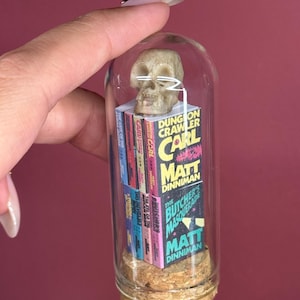 May include: A miniature glass cloche with a cork base containing a stack of tiny books titled Dungeon Crawler Carl by Matt Dinniman, including The Butcher's Masquerade, topped with a small, off-white, realistic human skull replica.