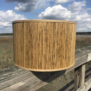 May include: A cylindrical lampshade made of vertical, natural-colored bamboo reeds. The lampshade is set against a backdrop of a wooden railing, marshland, and a cloudy blue sky. The lampshade has a light brown trim.