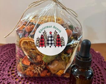 May include: A clear plastic bag filled with dried potpourri, including orange slices and pinecones, tied with twine. A circular label features the text Christmas Hearth and a graphic of five stylized trees. A small amber glass dropper bottle sits nearby.