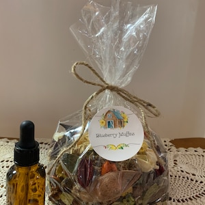 May include: A brown glass bottle with a dropper next to a clear cellophane bag tied with twine. The bag contains dried botanicals and a label that reads "Blueberry Muffins" with a barn illustration.