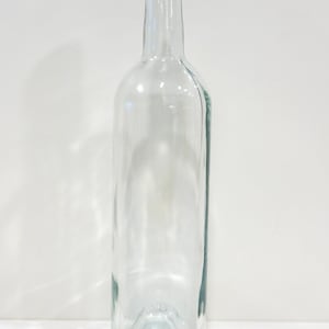 May include: A clear, empty glass bottle with a tall, slender neck and a threaded top. The bottle features a cylindrical body with a slightly concave base, showcasing a smooth, transparent surface throughout its entire structure.