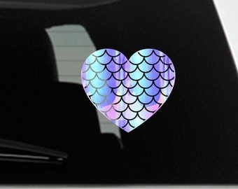 May include: A heart-shaped sticker with a mermaid scale pattern in iridescent blue, purple, and pink colors. The sticker is on a white car.
