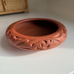 May include: A terracotta-colored, round bowl with carved floral and geometric patterns. The rim features small, evenly spaced indentations. The bowl is likely handmade and suitable for decorative or functional use.