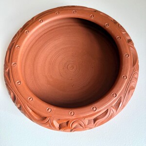 May include: A terracotta-colored, round ceramic bowl. The rim features a series of small, evenly spaced circular indentations and carved floral patterns. The bowl's interior is smooth, with concentric circles visible at the base.