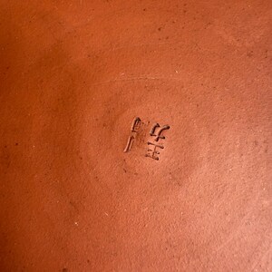 May include: Close-up of a terracotta-colored ceramic surface with stamped Japanese characters. The surface is a warm, earthy tone, and the characters are slightly darker, creating a subtle contrast. The texture appears smooth.